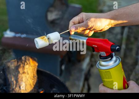 Toasting Marshmallows with a Portable Gas Torch. A hand toasts marshmallows on a wooden skewer with a portable gas torch beside an outdoor fire pit. Stock Photo