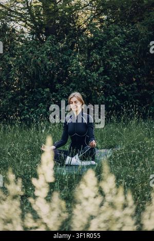 Young woman meditating outdoors on yoga mat. Self care routine, digital detox lifestyle, meditation for beginners. Stock Photo