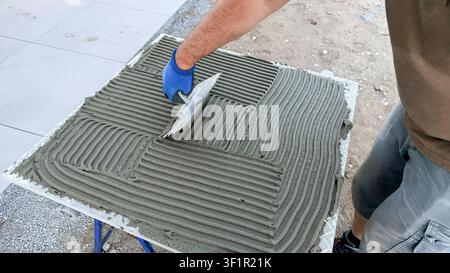 Construction worker applying cement mortar with trowel on a flat surface during flooring installation at a building site with tiles in the background Stock Photo