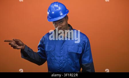 Man in blue hardhat and blue coveralls points finger to left, visible hand and finger, wearing safety helmet and work uniform in studio; focus guidanc Stock Photo
