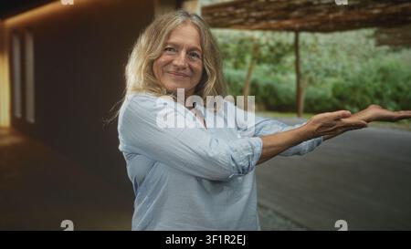 Senior woman with outstretched palms gesturing and smiling while presenting beside a wooden pergola building with greenery; contentment nature appreci Stock Photo