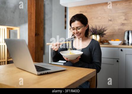 Happy Woman Takes A Working Break Eating Noodles With Laptop On The Kitchen Table Stock Photo