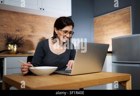 Smiling Woman Eating Noodles And Working On A Laptop At The Kitchen Table Stock Photo
