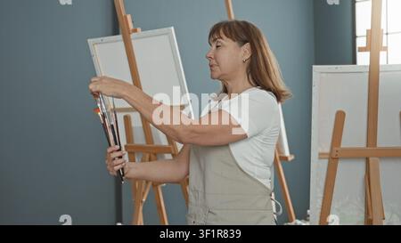 Woman painter selects paintbrushes and adjusts brush tips before blank canvas in studio; creative focus. Stock Photo