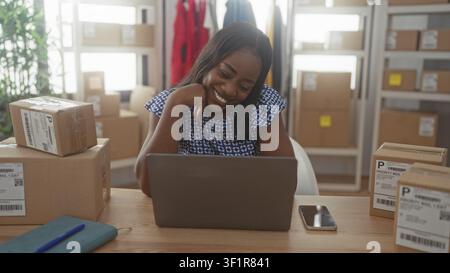 Woman at laptop resting chin on hand inside a packing building filled with boxes and shelves; entrepreneurial confidence. Stock Photo