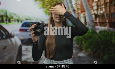 Woman adjusts dslr camera while checking settings on street with parked cars and trees; focus technique practice. Stock Photo
