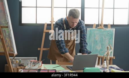 Man leans over laptop typing with hands on keyboard in art studio beside easel and paint brushes, mannequin and canvases; focused creativity. Stock Photo