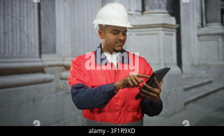 Hispanic man construction worker in hard hat points finger to tablet at building while checking plans in red jacket; confidence efficiency safety. Stock Photo