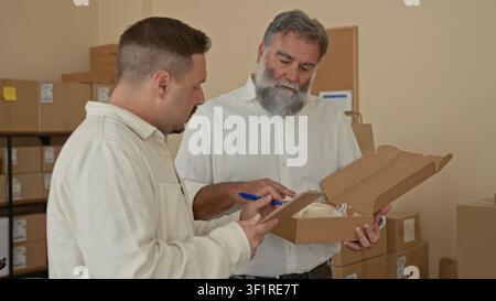 Two men inspecting a parcel, one writing on a clipboard while the other carefully holds an open box of packing filler amid stacked cartons in a storag Stock Photo