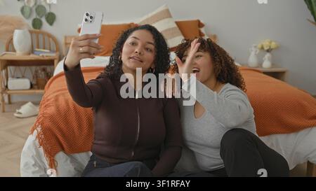 Woman holds smartphone aloft taking a selfie while companion rests her head on the other's shoulder in a cozy bedroom with orange bedding; joy friends Stock Photo