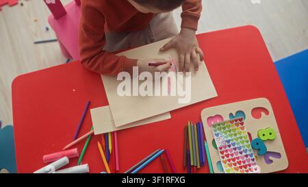 Boy tracing a hand and holding a red crayon while coloring on paper at a red kids table in a studio with stickers and colored pencils; concentration. Stock Photo