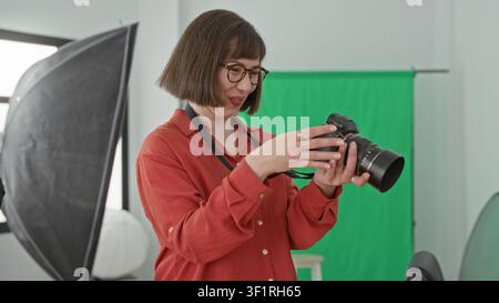 Woman photographer holding a dslr camera and checking images on the camera screen in a studio with green screen, softbox and stool; creative joy. Stock Photo