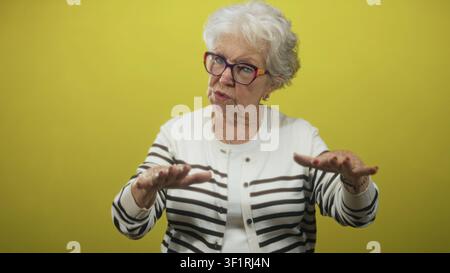 Senior woman with hands reaching forward gesturing, wearing striped cardigan and eyeglasses in studio; thoughtful reflection. Stock Photo