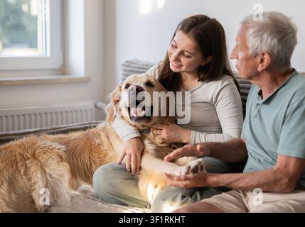 Happy Girl With Grandfather Playing With Golden Retriever Dog Sitting On A Sofa Stock Photo
