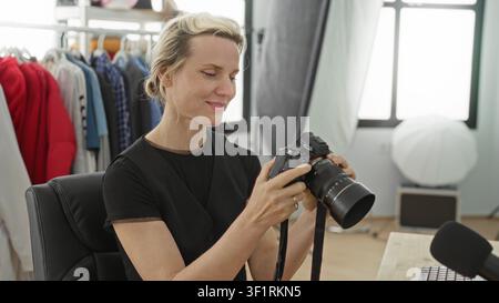 Woman adjusts camera lens and reviews images on computer in studio with wardrobe rack and softbox lighting; focused concentration. Stock Photo