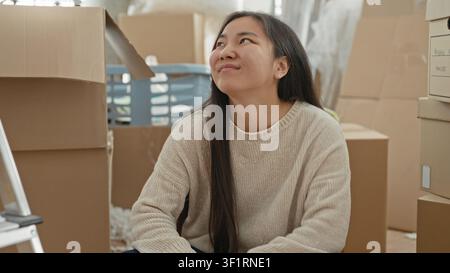 Woman sitting amid stacked cardboard boxes and a ladder, smiling while packing in a building during relocation; contentment moving day. Stock Photo