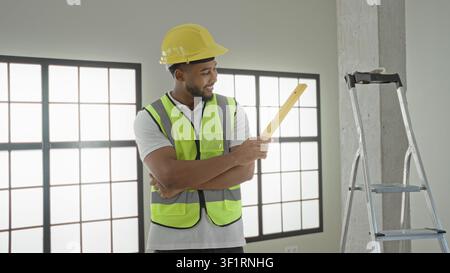 Man builder in hardhat smiles and holds a yellow measuring level with arms crossed revealing forearm inside a building; confidence professionalism. Stock Photo