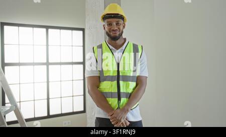 Man tipping hardhat with hand and smiling while wearing reflective vest in a building; confidence professionalism. Stock Photo