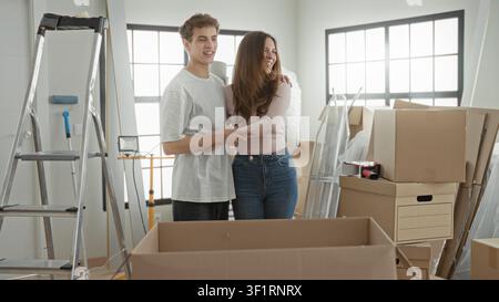 Woman and man hug among stacked cardboard boxes and ladder in an apartment while unpacking, woman gestures toward window and points; hopeful beginning Stock Photo