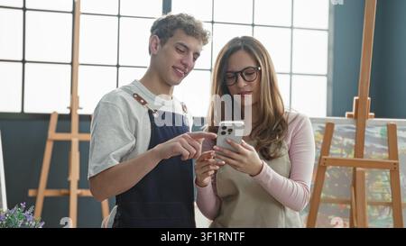 Man painter points finger to smartphone held by woman in studio while both wear aprons and stand by easels; creative collaboration joy. Stock Photo