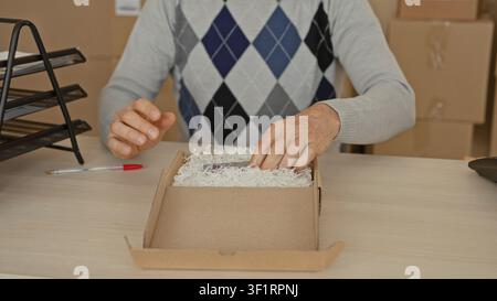 Man taps smartphone while packing a cardboard box with shredded paper in a building; small business optimism. Stock Photo