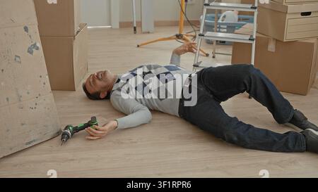Man lying unconscious on floor with hand near cordless drill beside stacked cardboard boxes and ladder in building interior new home moving scene; acc Stock Photo