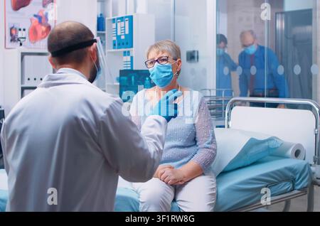 Old senior woman having a medical consultation during public health pandemic, wearing a mask and doctor in protective wear. Modern hospital or private clinic. Healthcare system. Stock Photo