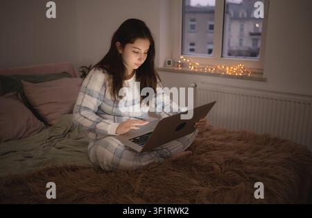 Girl sits on bed and uses laptop to chatting with friends in social media Stock Photo
