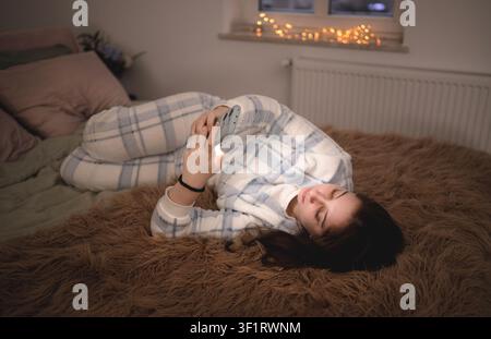 Girl lies on furry blanket in her room, focused on her mobile phone, typing messages in social media Stock Photo