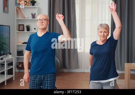 Happy senior couple exercises together on a yoga mat, practicing home workout, fitness and healthy active aging during retirement in the living room. Stock Photo