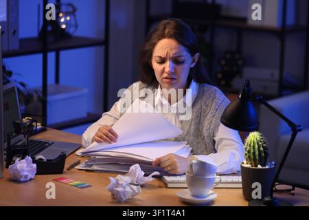 Overwhelmed young woman working late at night, stressed with a large stack of documents and crumpled paper on her desk Stock Photo