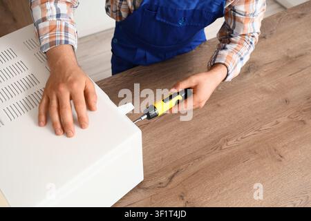 Repairman in blue uniform and plaid shirt fixing a white electronic device with a screwdriver on a wooden table, He uses a yellow and black screwdrive Stock Photo