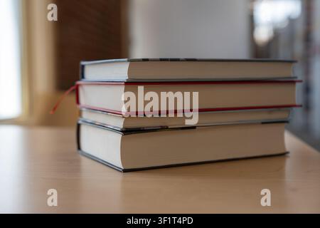 A close-up horizontal shot of a stack of three thick hardcover books lying on a light wooden table. The middle book features a distinct red edge or bo Stock Photo