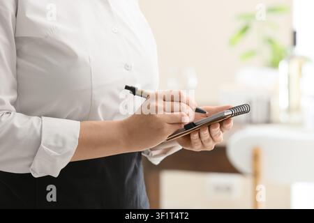 Waitress or server taking an order with a pen and notepad in a restaurant or cafe setting, the individual wears a white shirt and a black apron, indic Stock Photo