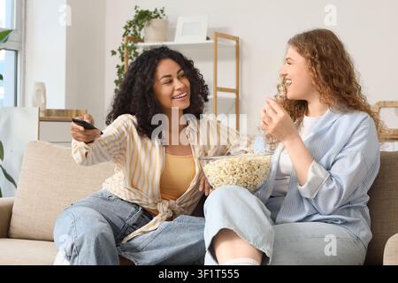 Two happy young women watching television and eating popcorn on a comfortable sofa at home, They are enjoying leisure time together, laughing and smil Stock Photo