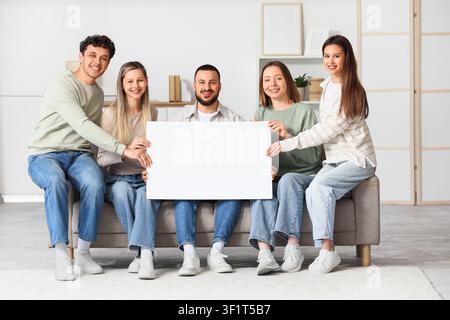 Five smiling young adults holding a blank white sign together while sitting on a sofa in a bright living room Stock Photo