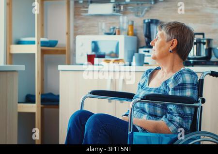 Handicapped senior woman in a wheelchair looks through a kitchen window alone, reflecting solitude, injury recovery and emotional sadness, during a focused consultation with clinical context. Stock Photo
