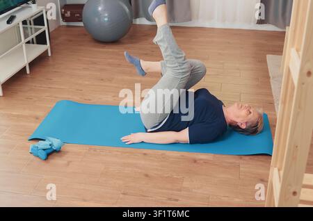 Elderly woman exercises while sitting on a yoga mat in the living room, supporting senior fitness, home training and healthy aging, as part of a realistic everyday lifestyle moment. Stock Photo