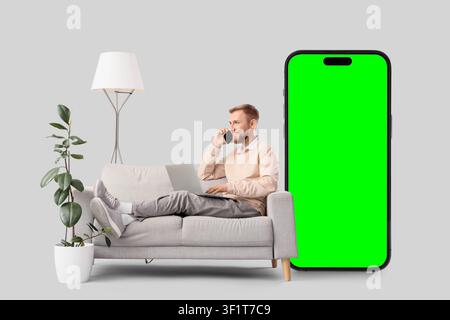 Man on sofa talking on phone and using laptop next to large smartphone with green screen remote work and communication, technology Stock Photo