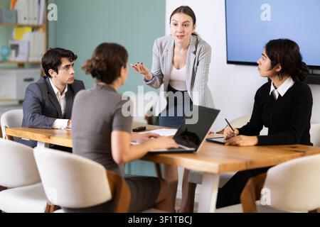 Confident young businesswoman standing near table where sitting colleagues talking and discussing ideas in office Stock Photo