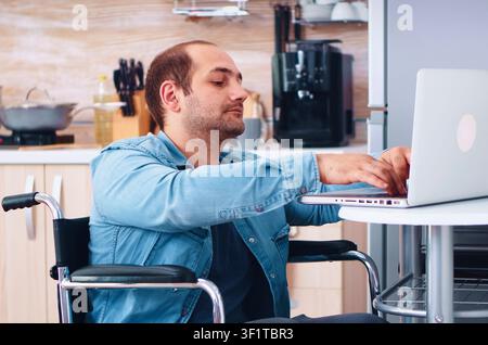 Man with special needs works on a laptop in the kitchen, combining remote business tasks with accessible home life, with the kitchen environment showing accessibility, support and daily living. Stock Photo