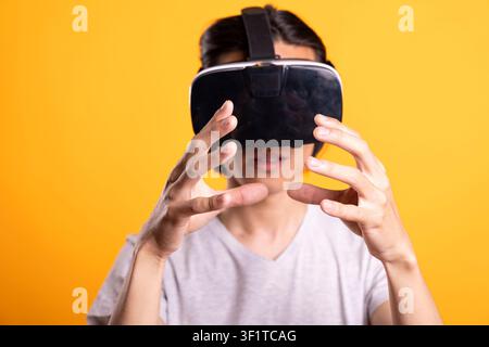 Asian man wears VR headset and reaches forward with hands, eating virtual burger. Person immersed in 3D digital world, enjoys playful augmented food interaction in studio with isolated background. Stock Photo