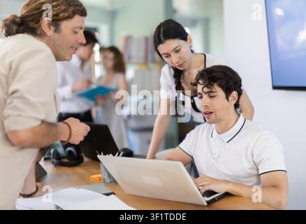 Several employees sit at desk during working day, working with laptop and paper documents. Stock Photo