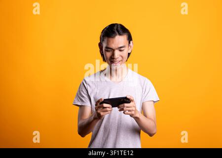 Happy male individual plays video games on mobile device with focus and excitement. Modern gamer enjoying interactive digital entertainment and online gaming on smartphone in yellow background studio. Stock Photo