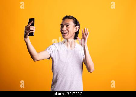 Cheerful Asian man holds smartphone and waves while smiling in front of yellow background. Friendly young adult enjoys video call, showing online communication, connection and positive lifestyle. Stock Photo