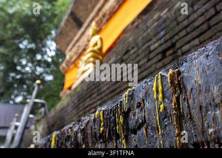 A burned wax drips down outdoor stone ledge near robe draped golden buddha statue, creating textured yellow streaks and sombre ritual atmosphere in te Stock Photo