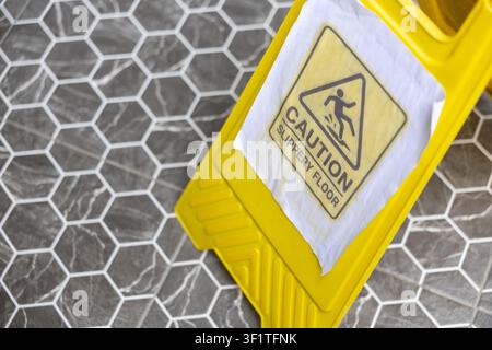 A caution wet floor sign on wet hexagon tile floor warning pedestrians to avoid slipping and showing bright yellow safety marker Stock Photo