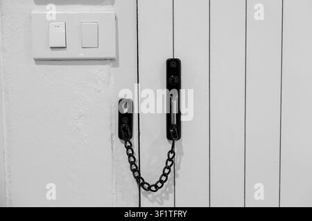 A door chain lock on white wooden door with vertical panels and light switchplate, minimal monochrome interior scene showing security detail and contr Stock Photo