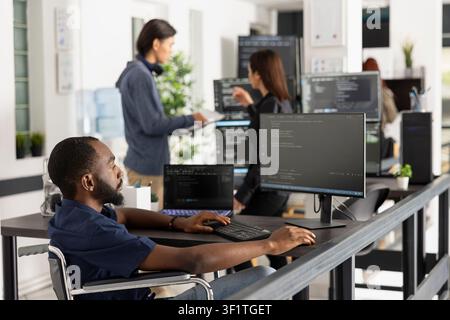 Black woman programmer works at desktop computer in modern startup office workspace. Focused developer in wheelchair represents innovation, accessibility, and diversity in the technology sector. Stock Photo