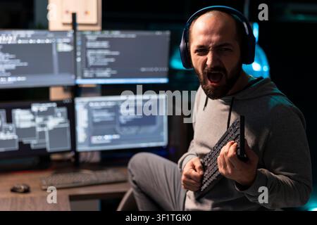 Tired and cheerful developer relaxes after coding, holding keyboard in humorous pose. Male programmer taking playful break from work, pretending to play music with computer keyboard. Stock Photo